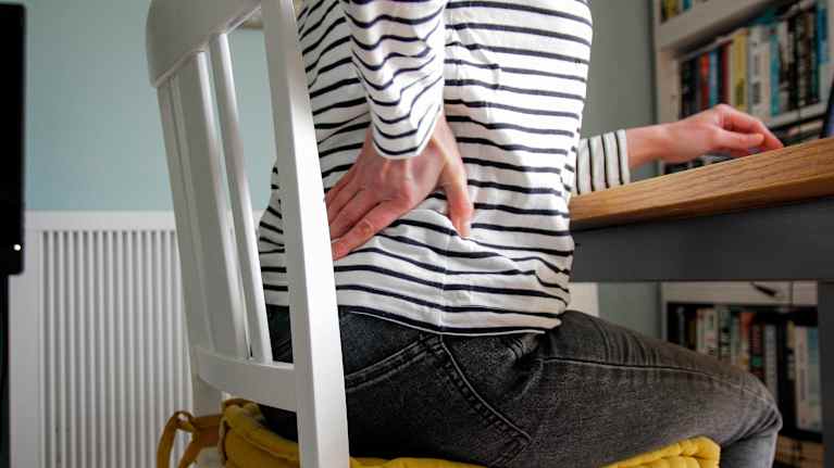 Ihminen istuu työpöydän ääressä ja pitelee kädellä alaselkäänsä.