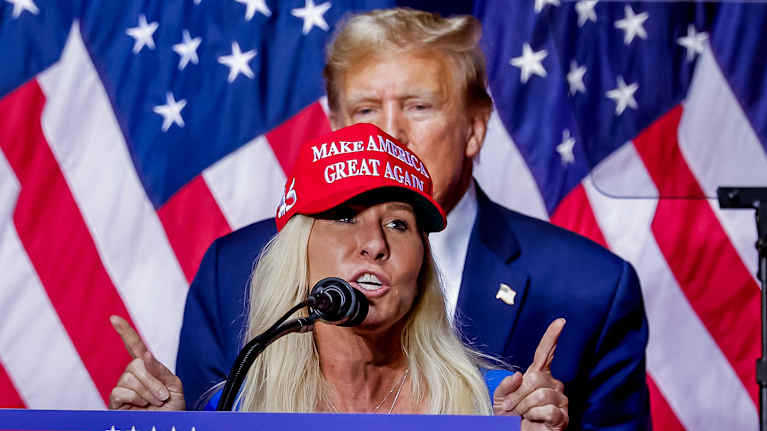 Marjorie Taylor Greene puhuu punaisessa Make America Great -lippiksessä puhujanpöntön takana. Donald Trump seisoo hänen takanaan.