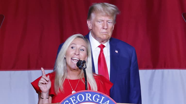 Marjorie Taylor Greene puhuu punaisessa leningissä puhujanpöntössä. Takana on Donald Trump sinisessä puvussa ja kravatissa. Pöntössä lukee Georgia.