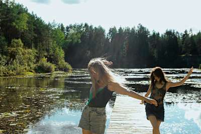 Kaksi nuorta tyttöä farkkushortseissa ja hihattomissa t-paidoissa kävelee tanssimaisin askelin laiturilla kesäisessä järvimaisemassa.