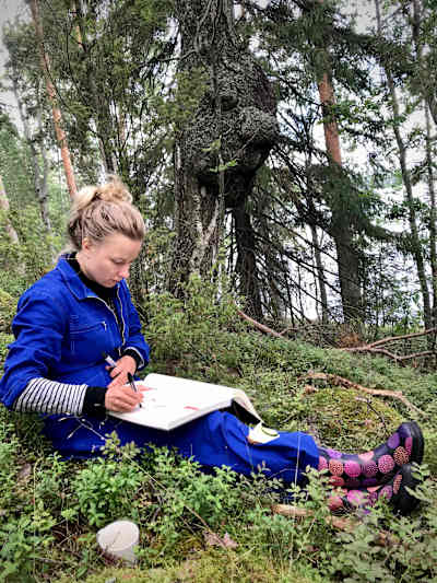 Nainen (näyttelijä Henna Tanskanen) sinisessä haalarissa ja kirjavissa kumisaappaissa nojaa suureen kiveen kesäisessä metsässä ja piirtää isoon kovakantiseen lehtiöön.