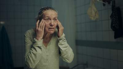 Keski-ikäinen nainen (näyttelijä Riitta Havukainen) suihkumyssyssä tarkastelee silmäkulmiensa ryppyjä kylpyhuoneen peilistä.