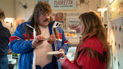 Tukeva, viiksekäs, pitkätukkainen mies (näyttelijä Hannu-Pekka Björkman) pitelee hillomunkkia kahvilan pihdeissä ja katsoo nuorempaa naista (näyttelijä Oona Airola), joka puhuttelee häntä.