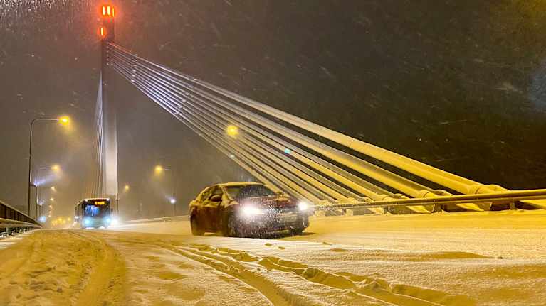 Talvinen iltatunnelma lumisateen keskellä Jätkänkynttilän siltaa Rovaniemellä; auto lipuu hiljalleen eteenpäin lumipeitteisellä tiellä