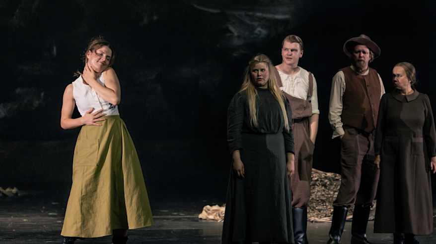 Nuori nainen pitkässä hameessa. Neljä henkilöä katselee häntä sivussa.