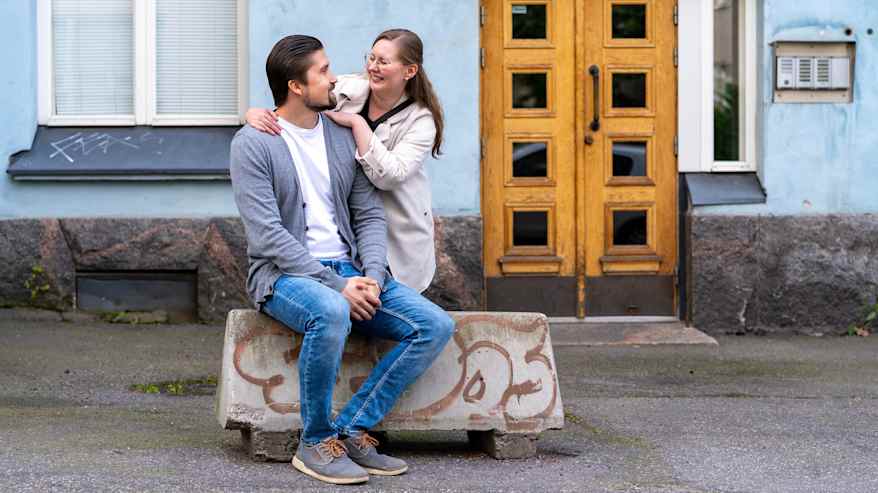 Mikko ja Hanna Poikkilehto istuvat yhdessä kadulla betoniporsaan päällä, taustanaan vanha rakennus puisella ulko-ovella.