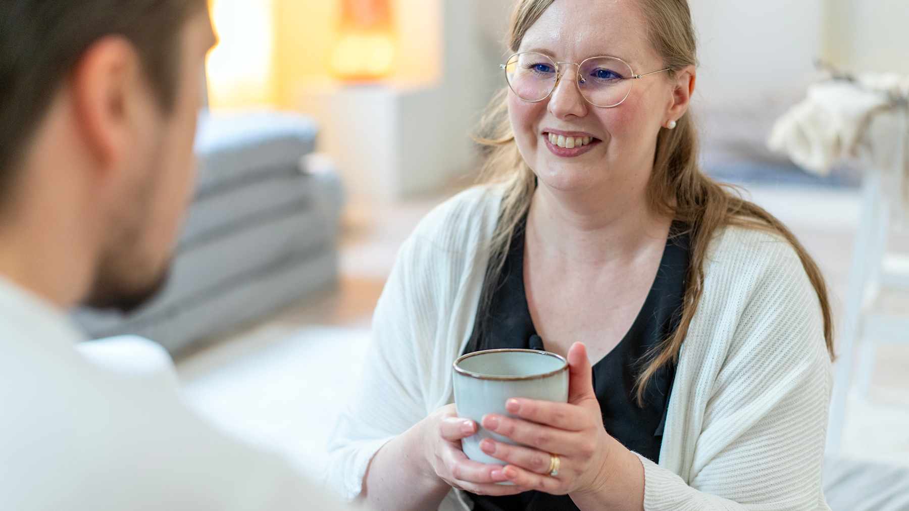 Hanna Poikkilehto pitää kädessään mukia ja näyttää keskustelevan toisen henkilön kanssa kodikkaassa ympäristössä.