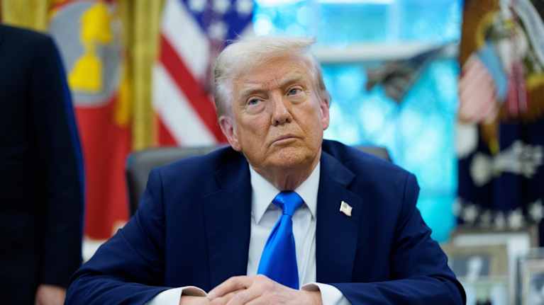 Harmaapäinen mies sinisessä puvussa työpöydän takana. 