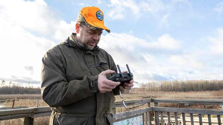 Henkilö ohjaa dronea ulkoilmassa lintutornilta käsin, taustalla avautuu syksyinen maisema.