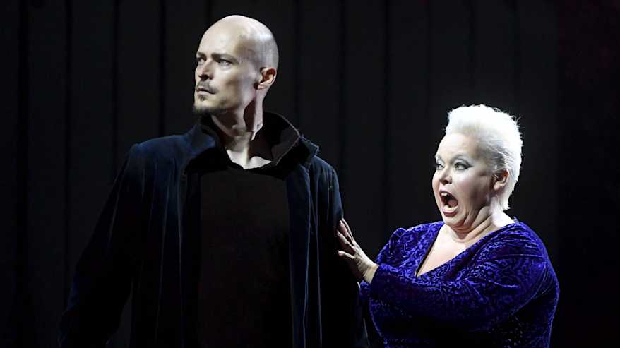 Johanna Rusanen-Kartano pitää toiselle kädellä Ville Rusasen olkapäästä ja laulaa suu isosti auki sinertävässä mekossa.