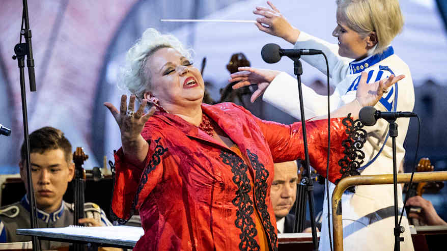 Johanna Rusanen-Kartano laulaa kädet levällään punaisessa asussa lavalla. Taustalla soittokunta.