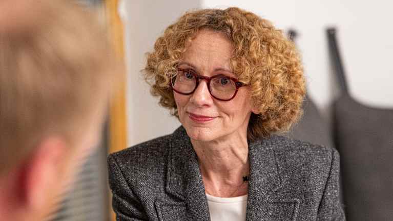 Kuvassa henkilö, jolla on kihara hiustyyli ja harmaa pikkutakki, keskustelemassa toisen henkilön kanssa sisätiloissa.