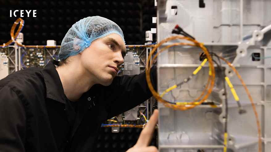 Hiussuojaverkkoon ja suojatakkiin pukeutunut mieshenkilö tarkistaa metallista laatikkoa, jonka kyljessä näkyy johtoja.