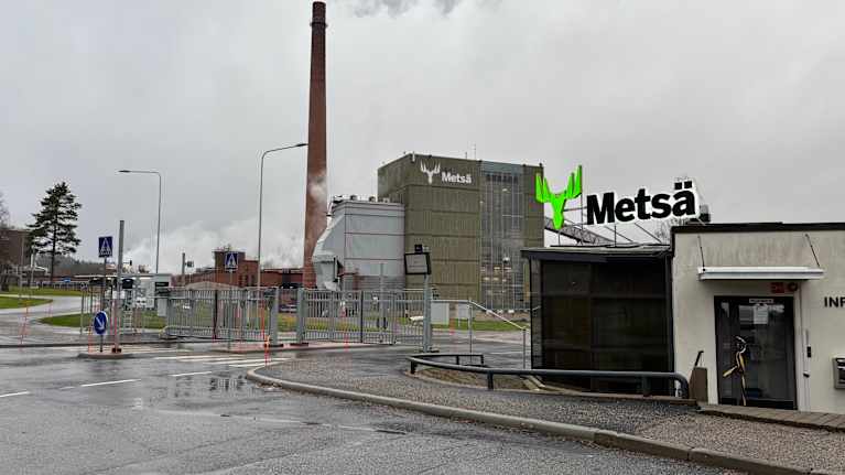Metsä Groupin teollisuuslaitos harmaana syyspäivänä. Tehdasrakennuksesta nousee savua taivaalle, ja taustalla kohoaa korkea savupiippu.