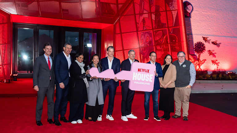 Ryhmä ihmisiä seisoo punaiseksi valaistun sisäänkäynnin edessä kädessään iso vaaleanpunainen avain, jossa lukee Netflix House.