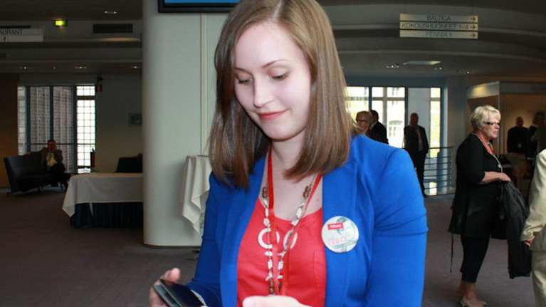 Sanna Marin vuonna 2012 kädessään kaksi kännykkää ja päällään sininen jakkutakki ja punainen paita.