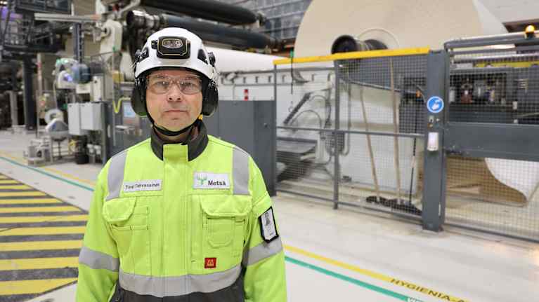 Metan työntekijä seisoo tehtaassa kartonkikoneen edessä, pukeutuneena huomioväriseen työasuun suojavarusteineen.