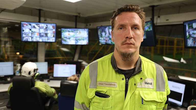 Mies seisoo työvaatteissa valvontakeskuksessa monitorien edessä. Taustalla työkaverit keskittyvät monitorien tarkkailuun.