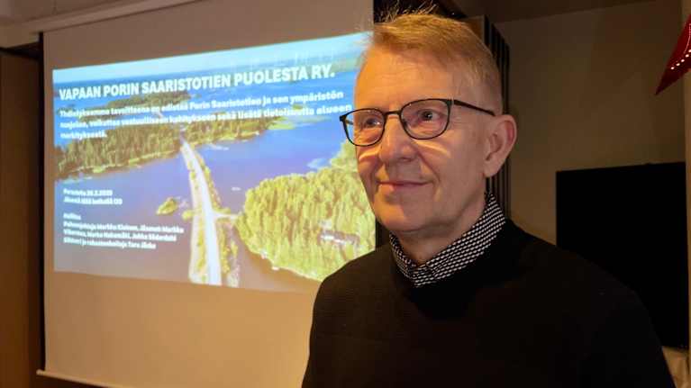 Mies seisoo huoneessa, taustalla diaesityksessä kuva Porin Saaristotiestä.