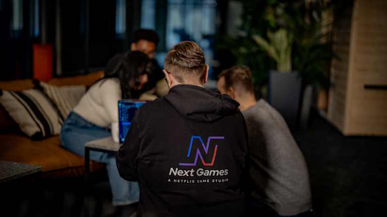 Next Games -peliyhtiön työntekijöitä selkä kameraan päin. Etualalla olevalla huppari, jossa takana Next Gamesin violetinsininen logo.