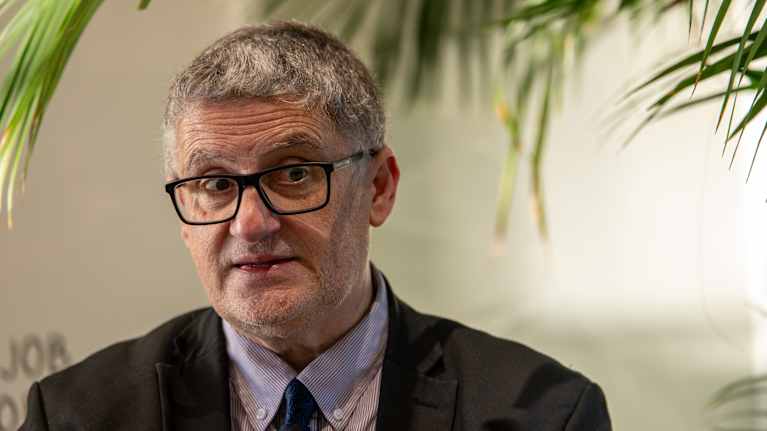 Henkilö keskustelee vilkkaasti sisätilassa, ympäröitynä vihreillä kasveilla. Hänellä on yllään tumma puku ja solmio.