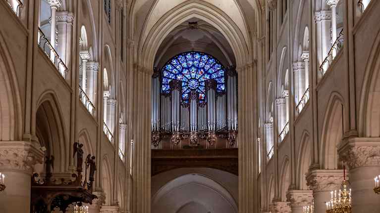 Notre Damen katedraali. Ihmisiä istuu käytävän molemmin puolin. Korkeassa huoneessa käytävän päässä urkuparvi, koristeellisen ikkunan alla.