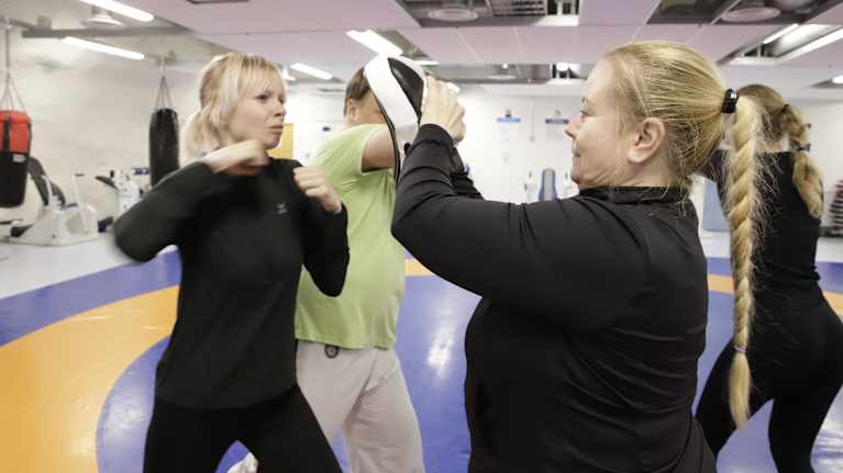 Nuori nainen lyö nyrkillä toisen naisen pitämää pehmustetta.