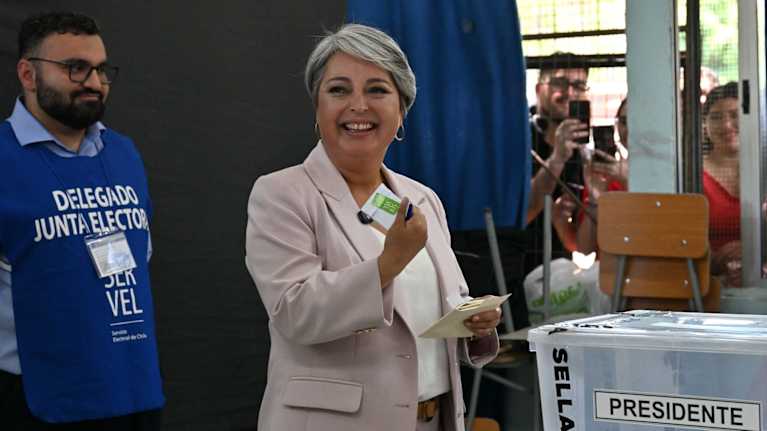 Jakkupukuun pukeutunut hymyilevä nainen pitelee käsissään äänestyslipukkeita äänestyspaikalla.