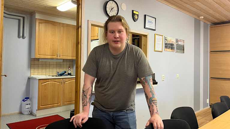 Mies seisoo toimistotilassa nojaten pöytään, hänen ympärillään on tuoleja.