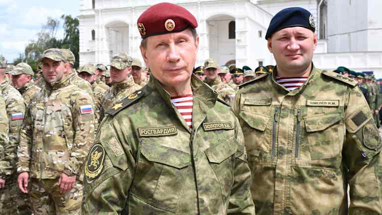 Zolotov seisoo maastokuvioidussa univormussa ja punaisessa baretissa kansalliskaartin joukkojen edessä. Taustalla on valkoisen kirkon julkisivu.