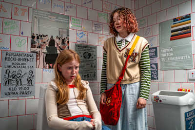 Kaksi tyttöä koulun vessassa. Punahiuksinen tyttö istuu pytyn kannen päällä ja näyttää huolestuneelta. Kiharahiuksinen tyttö seisoo vieressä.