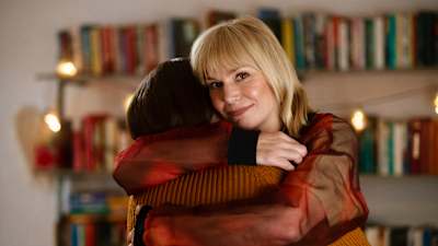 Lämminsävyisessä kuvassa toimittaja Eva Kela halaa naista ja katsoo hänen olkansa yli kameraan.