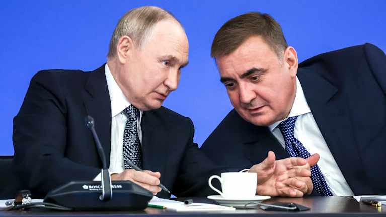 Putin ja Djumin istuvat mustissa puvuissa pöydän takana. Putin puhuu puoleensa kumartuneelle Djuminille kynä oikeassa kädessään,