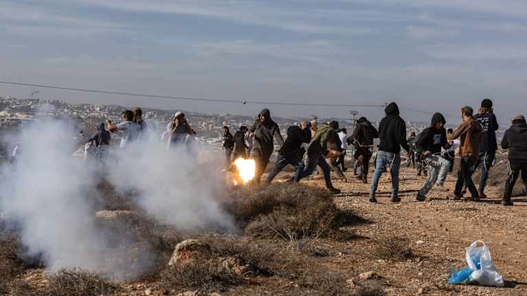 Huppareihin pukeutuneita nuoria miehiä kääntyy pakenemaan samalla, kun vieressä leimahtaa savukranaatti.
