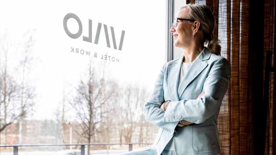 Tiina Laisi-Puheloinen seisoo sinisessä housupuvussa lenkkarit jalassa. ikkunan edessä.