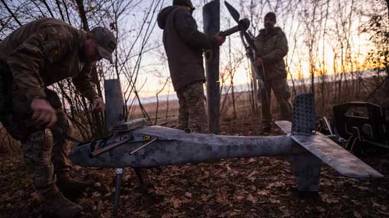 Kolme maastopukuista miestä valmistelee kiinteäsiipistä droonia lentoon metsänreunassa.