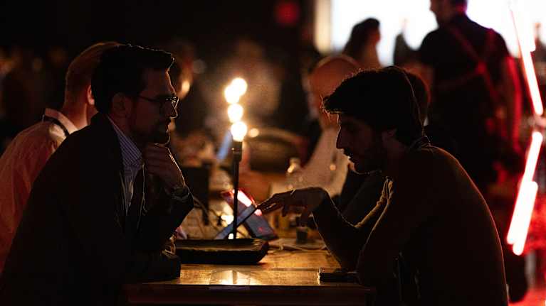 Pitkän pöydän ääressä ihmisiä keskustelemassa hämärässä valaistuksessa.