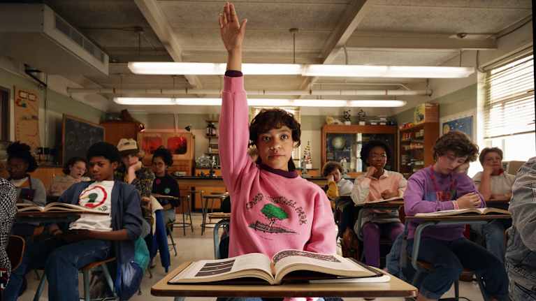 Priah Ferguson Erica Sinclairina sarjassa Stranger Things. Näyttelijä on luokkahuoneessa ja viittaa.