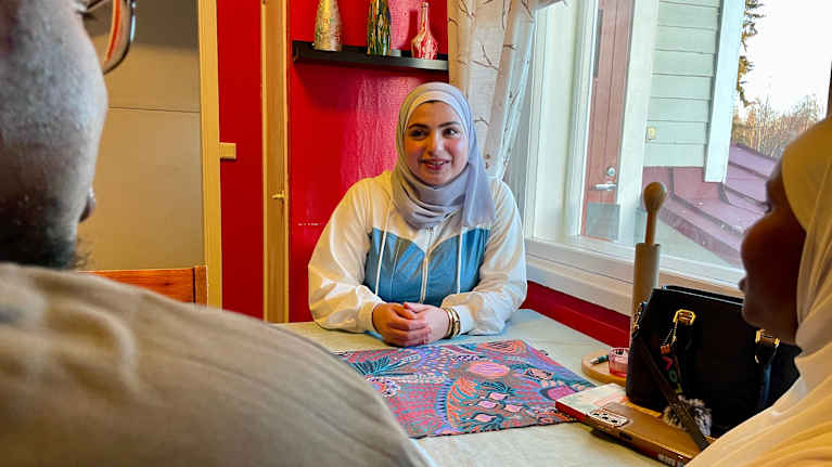 Huivipäinen nainen istuu pöydän ääressä. Hänen kätensä ovat levollisesti pöydällä.