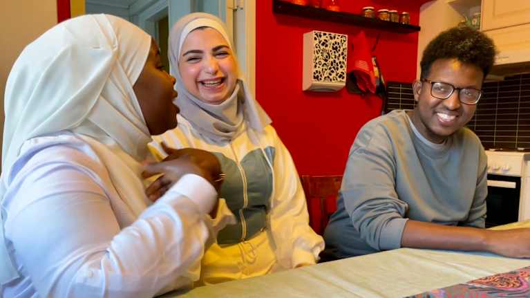 Kaksi huivipäistä naista istuu miehen kanssa pöydän ääressä. Toinen naisista selittää iloisesti ja toinen nainen ja mies nauravat.