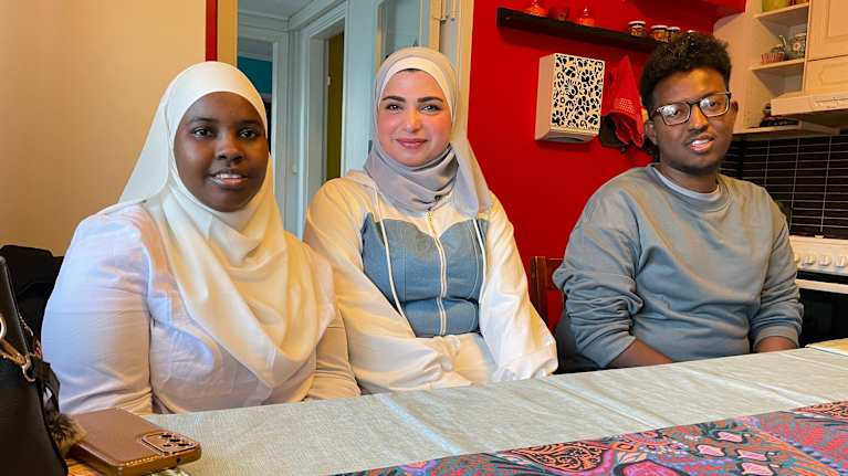 Kaksi huivipäistä naista ja mies istuvat rivissä pöydän ääressä. He katsovat kohti ja ilme on rento.