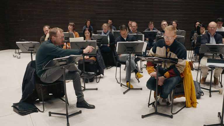 Ihmisiä istuu huoneessa nuottitelineiden takana. Etualalla kaksi miestä, joista toinen sanoo jotain toiselle.