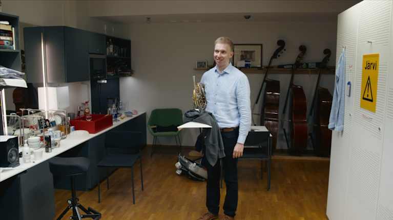 Hymyilevä mies seisoo huoneessa kädessään käyträtorvi. Taustalla rivi kontrabassoja.