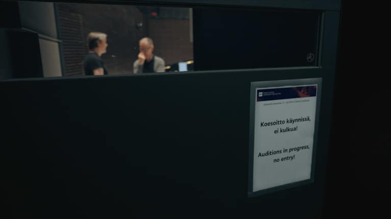 Ovessa on kyltti, jossa lukee "Koesoittokäynnissä, ei kulkua!". Ovessa olevan ikkunan läpi näkyy kaksi miestä, jotka keskustelevat.