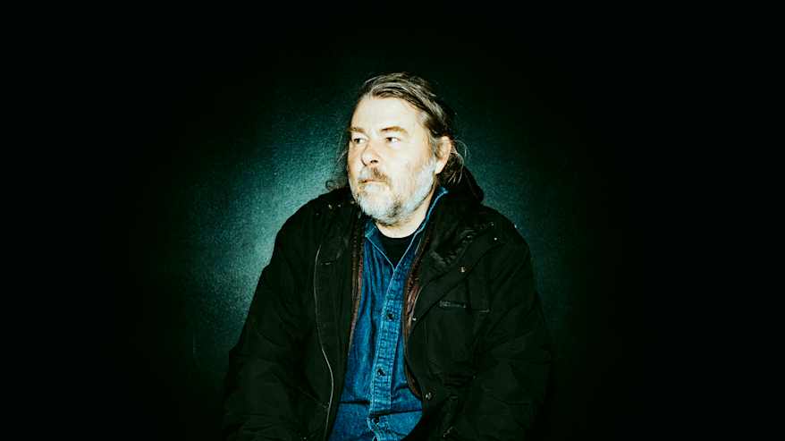 Henkilö on pukeutunut tummiin vaatteisiin ja istuu vasten tummaa taustaa.