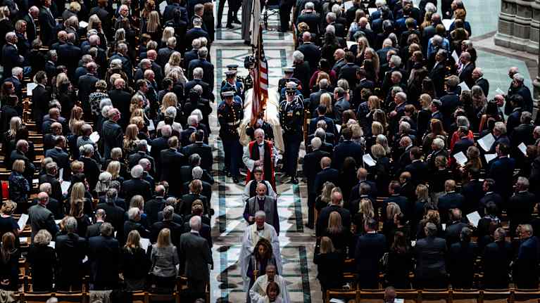 Kirkkosali ylhäältä päin kuvattuna, paljon ihmisiä, keskikäytävällä valkoisiin kaapuihin  pukeutuneita pappeja, sotilaat kantavat arkkua.