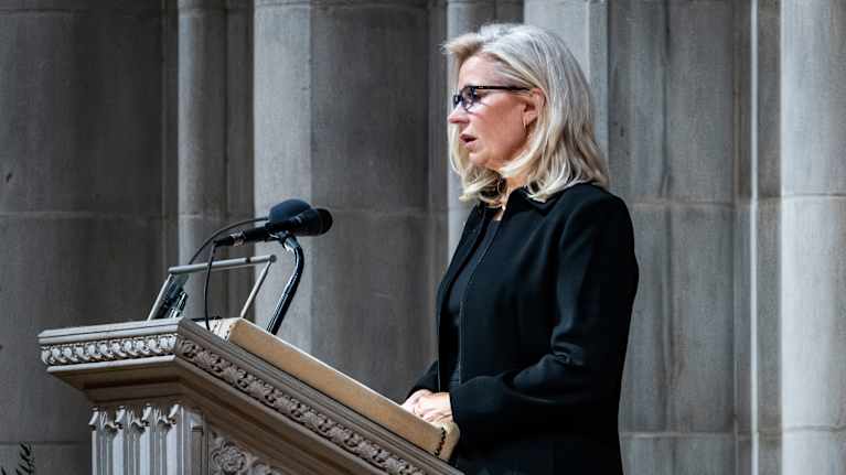 Mustaan jakkuun ja puseroon pukeutunut nainen seisoo puhujapöntössä, taustalla marmorilta näyttäviä pylväitä.