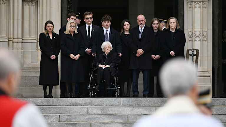 Mustiin pukeutuneet henkilöt seisovat kirkon portailla. Edustalla epätarkasti kuvattuna kaksi pappia.