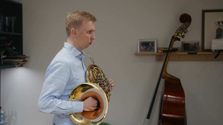 Mies seisoo käyrätorvi kädessään, toinen käsi on torvan sisällä ja toinen läpillä. Ilme on keskittynyt.