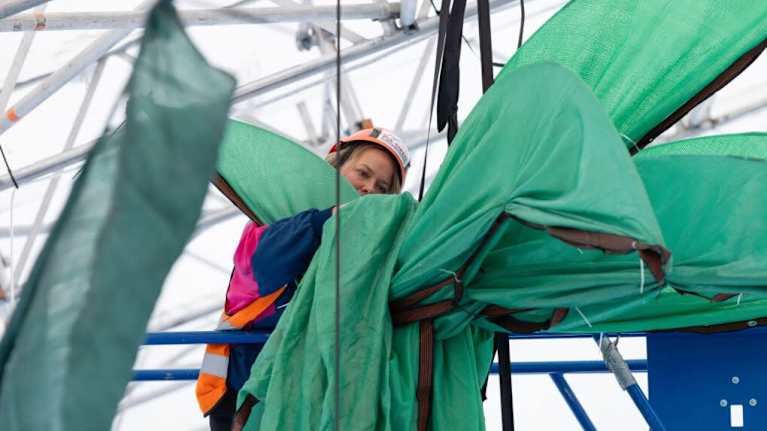 Nainen asentaa suuria vihreitä kankaita tai rakenteita korkealla metallikehikon sisällä. 